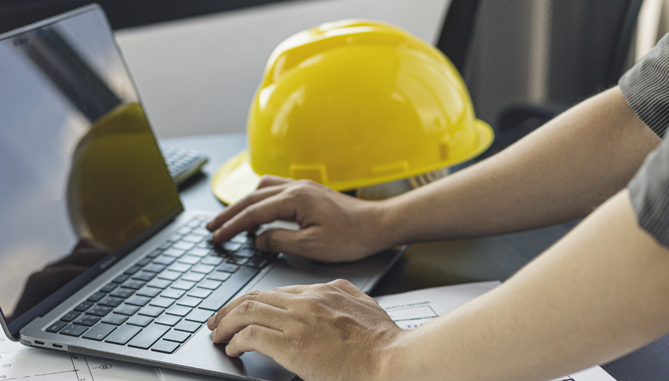 An image of someone resting their hands on the keyboard of a laptop computer with a construction-type hard hat resting on a table next to the laptop